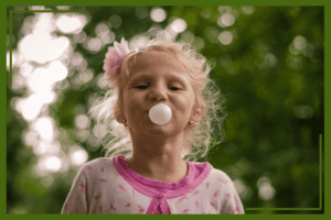 A young girl with curly blonde hair blows a bubble gum bubble, wearing a pink cardigan outdoors with a blurred green background.