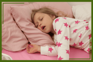 A young girl is sleeping on a pink bed with her mouth slightly open. She is wearing white pajamas with pink star patterns and resting her head on a soft pink pillow.