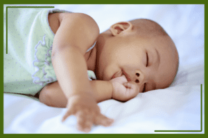 A sleeping baby in a light green outfit peacefully sucking their thumb while lying on a white bed.