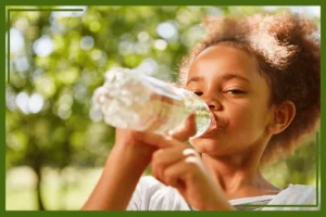 A young child drinking water from a clear plastic bottle outdoors with a blurred green background.